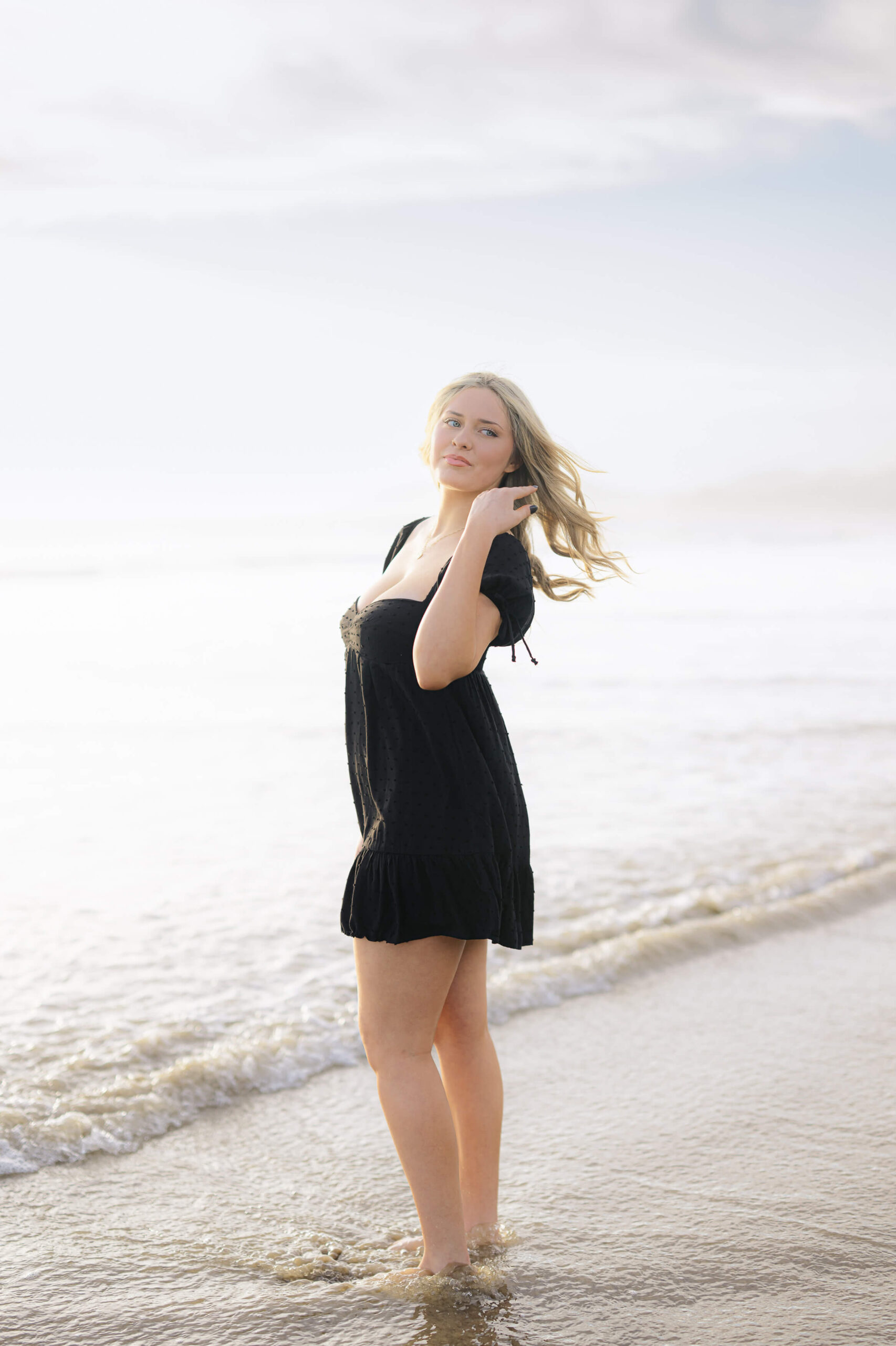 High school senior portrait at Pismo Beach during golden hour. A young woman in a black dress poses by the shoreline with ocean waves in the background, captured by San Luis Obispo photographer Rosa Clark.